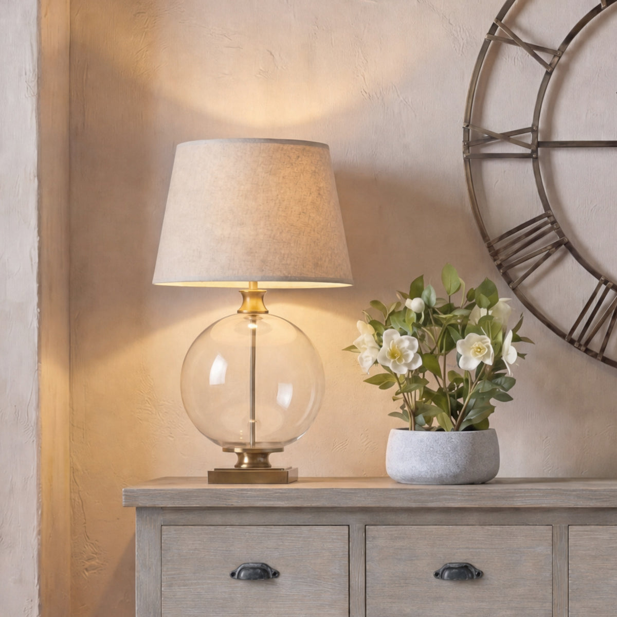Table lamp pictured lit on a grey timber sideboard beside white flowers in a grey pot, beneath part of a large open-face wall clock.