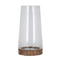 Candle lantern on a white background, with a tall clear open vessel tapering towards the top above a low round brown woven tray.