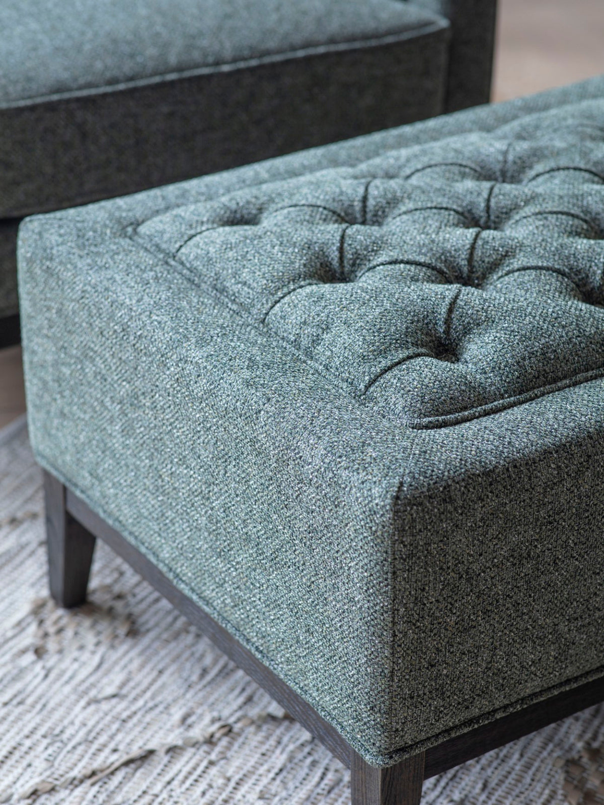 Close-up of the stool’s textured green and grey weave, deep button detailing and piped border, with part of an armchair behind.
