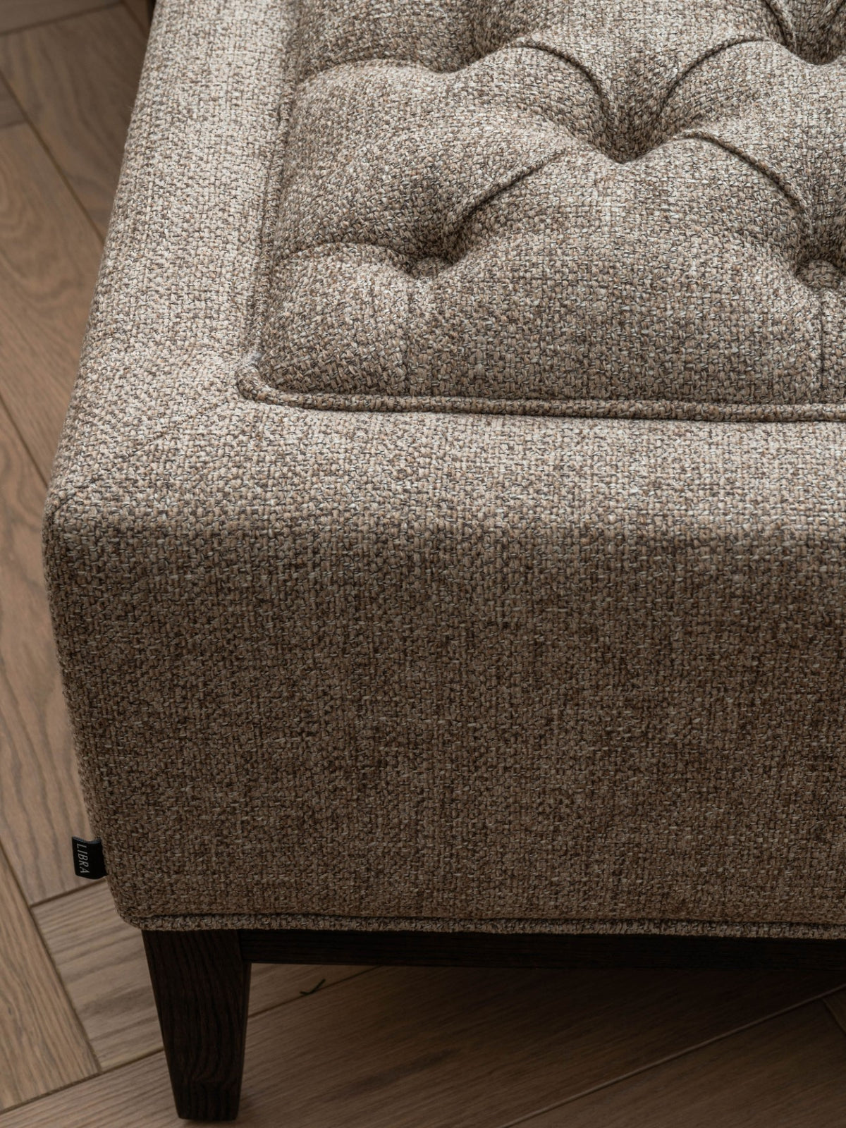 Close-up of the footstool's taupe and beige weave, deep fabric-covered buttons, piped top edge and dark wooden leg.