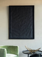 Black cobbled relief wall art on a pale wall above a green armchair and dark round table with a gold-toned bowl.