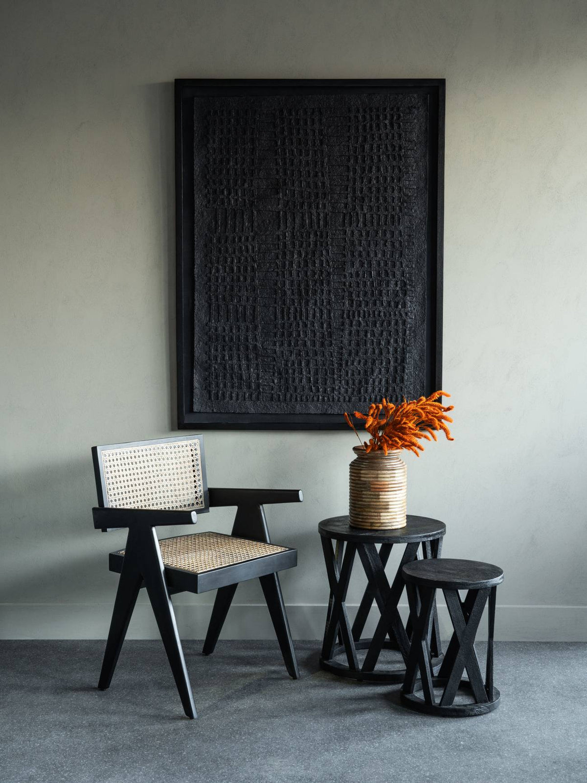 Cobbled relief wall art above a black cane chair and two black tables, with orange stems in a ribbed vase.