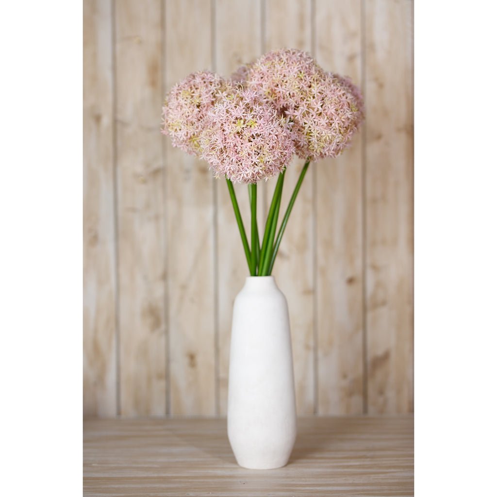 Pale lilac globe flowers in a tall white vase on a wooden surface, with pale wooden panelling behind.
