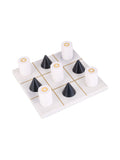 Angled view of a square white resin noughts-and-crosses board with a brass-tone grid, black conical pieces and white cylindrical pieces against a white background.
