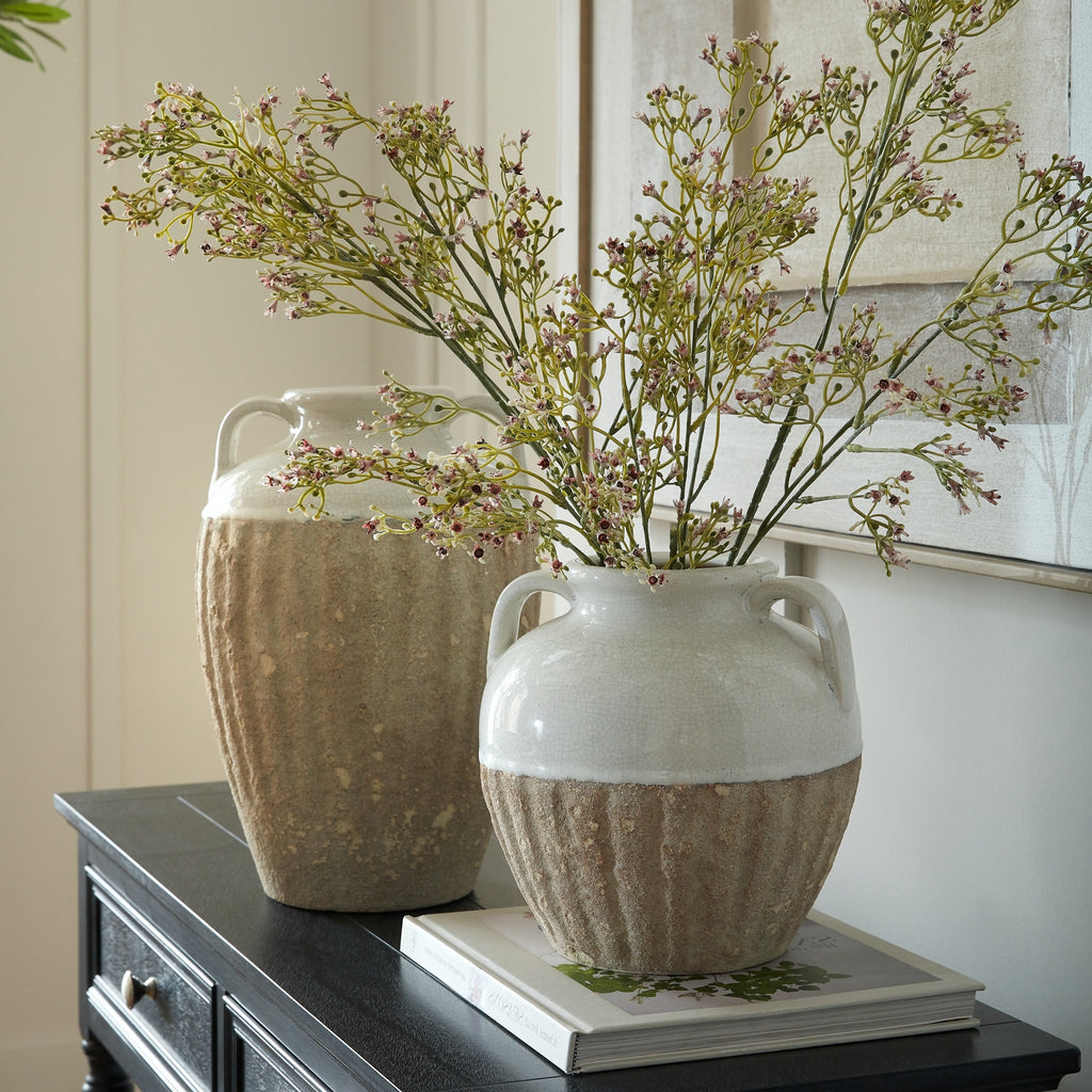Rounded shoulder-handled vase holding branching flower stems on a book, with a taller vase behind it on a dark console beneath artwork.
