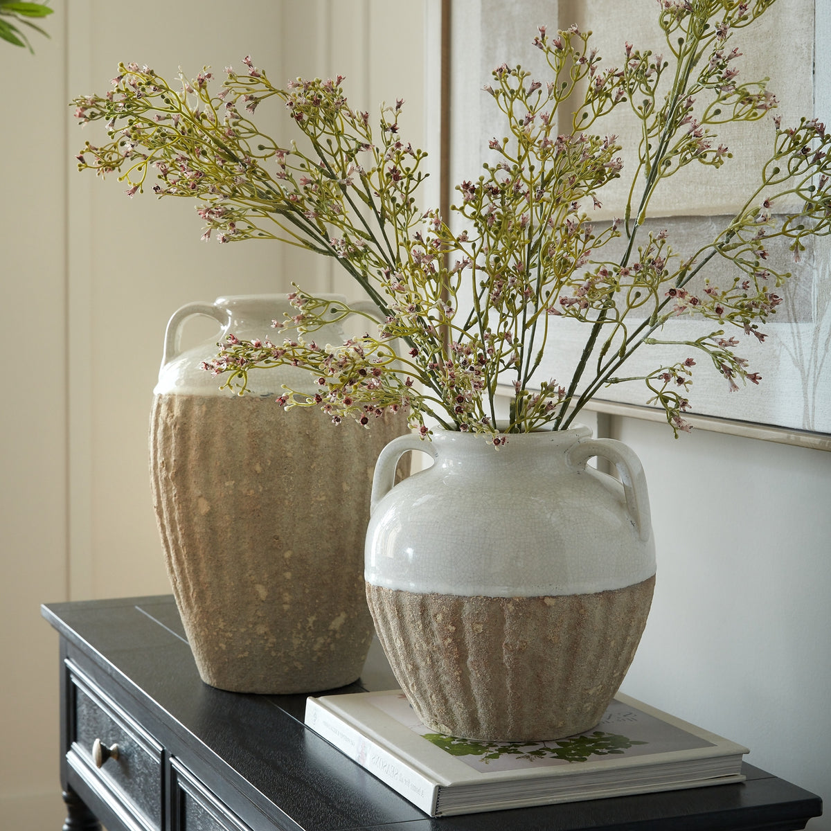 Rounded shoulder-handled vase holding branching flower stems on a book, with a taller vase behind it on a dark console beneath artwork.