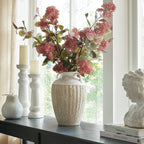 Two-handled vase filled with pink-red flowering stems on a dark console by a window, beside white candleholders and a bust on books.