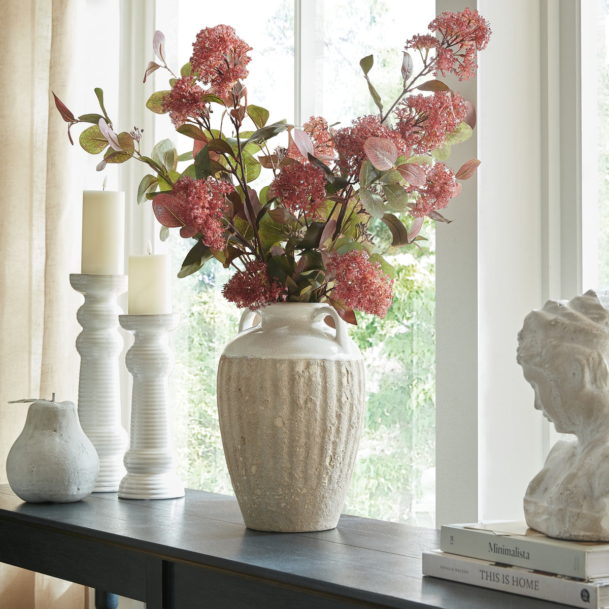 Two-handled vase filled with pink-red flowering stems on a dark console by a window, beside white candleholders and a bust on books.