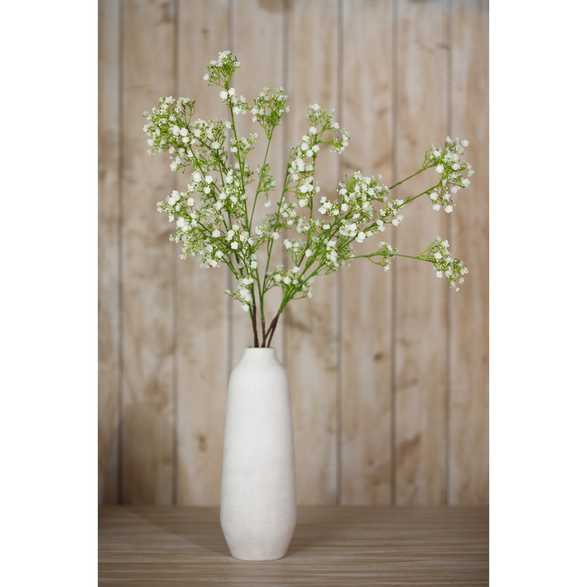 Cream artificial gypsophila arranged in a tall pale vase on a wooden surface in front of vertical wood panelling.