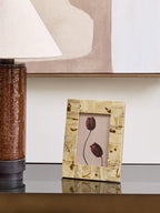 Cream patchwork frame holding a botanical print on a reflective console beside a brown lamp, with neutral artwork behind.