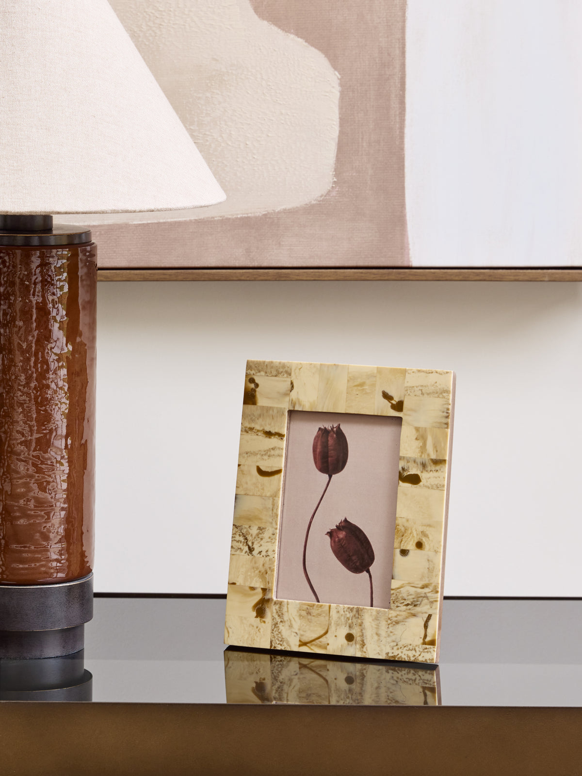 Cream patchwork frame holding a botanical print on a reflective console beside a brown lamp, with neutral artwork behind.