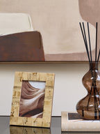 Cream patchwork frame displaying a sandy abstract photograph on a dark tabletop beside a brown glass reed diffuser and books.