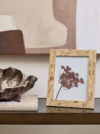 Cream patchwork frame holding a botanical image on a reflective console beside a sculptural bowl and book, beneath abstract artwork.