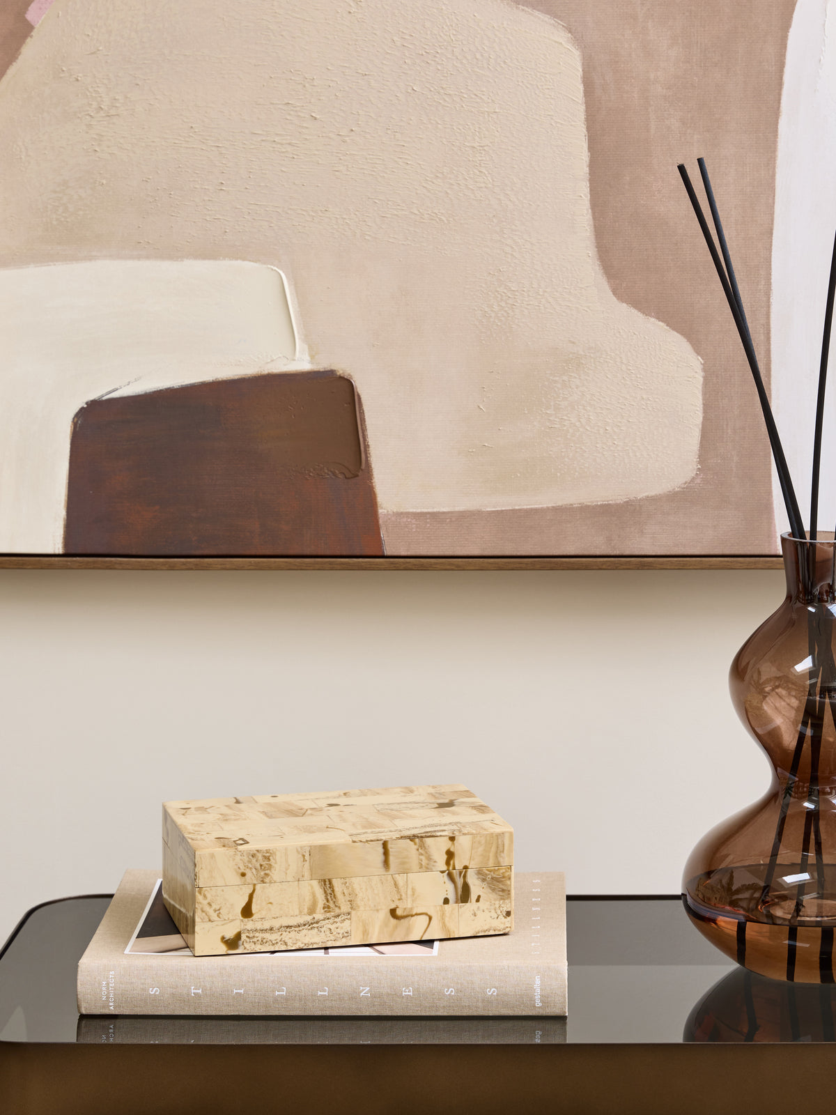 Cream patchwork storage box on a beige book, beside a brown glass reed diffuser and beneath neutral abstract artwork.