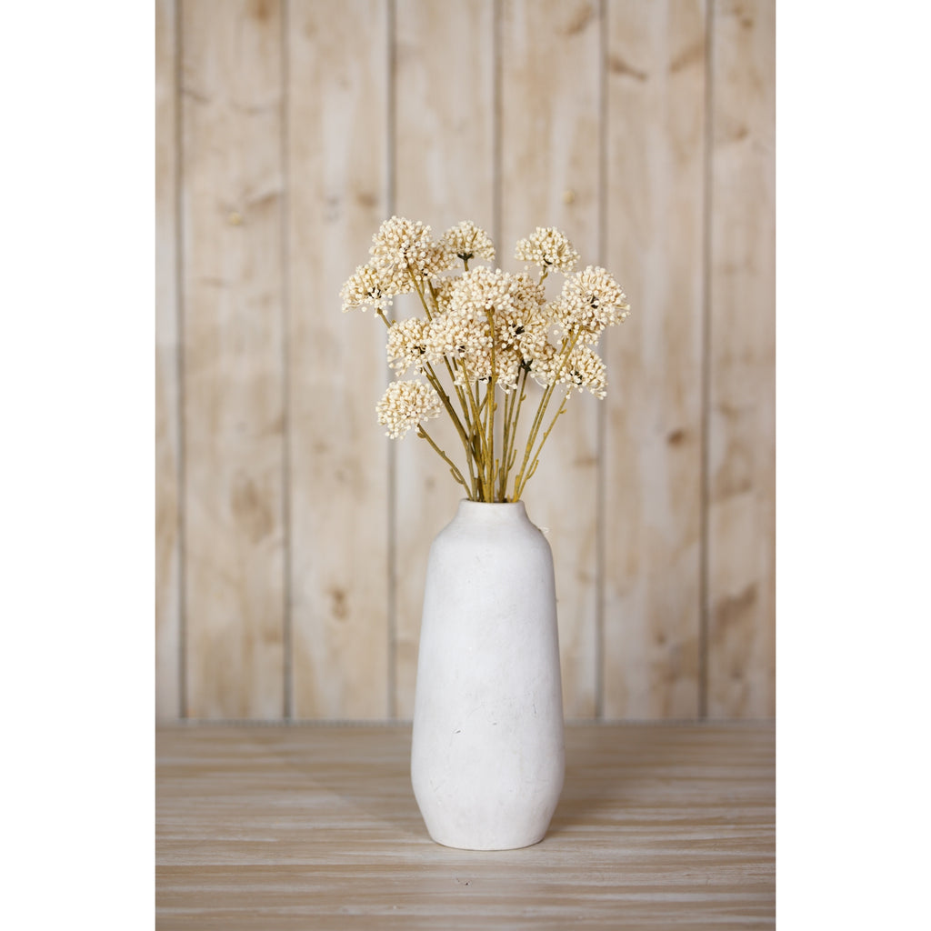 Cream botanical sprays displayed in a tall pale vase on a wooden surface in front of vertical wood panelling.