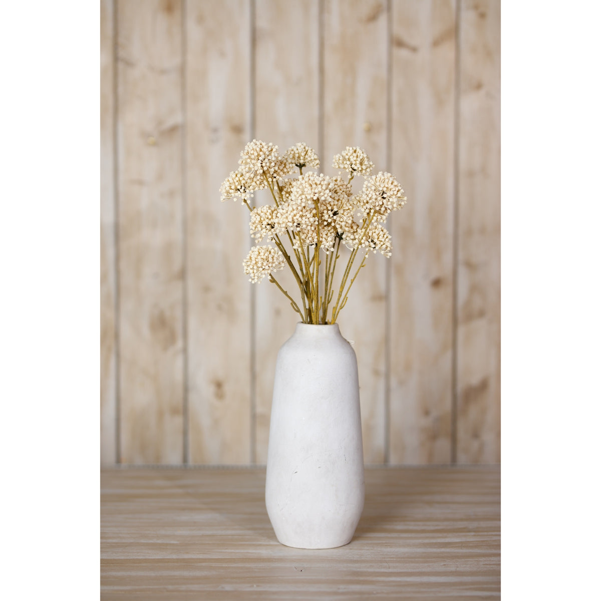 Cream botanical sprays displayed in a tall pale vase on a wooden surface in front of vertical wood panelling.