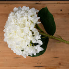 Close view of overlapping white petals with pale cream centres beside a dark green veined leaf, with the artificial stem lying on a wooden surface.