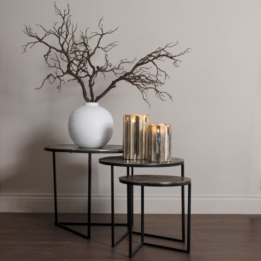 Bare artificial branches spreading from a rounded white vase on the tallest of three nesting tables, beside two glowing metallic candle holders.