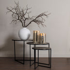 Bare artificial branches spreading from a rounded white vase on the tallest of three nesting tables, beside two glowing metallic candle holders.
