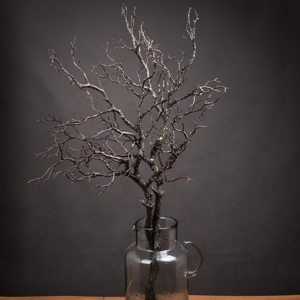 Speckled artificial branches arranged in a clear handled jug on a wooden surface against a dark wall.