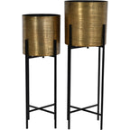 Two brass-coloured cylindrical planters at different heights on slim black stands with crossed lower supports.