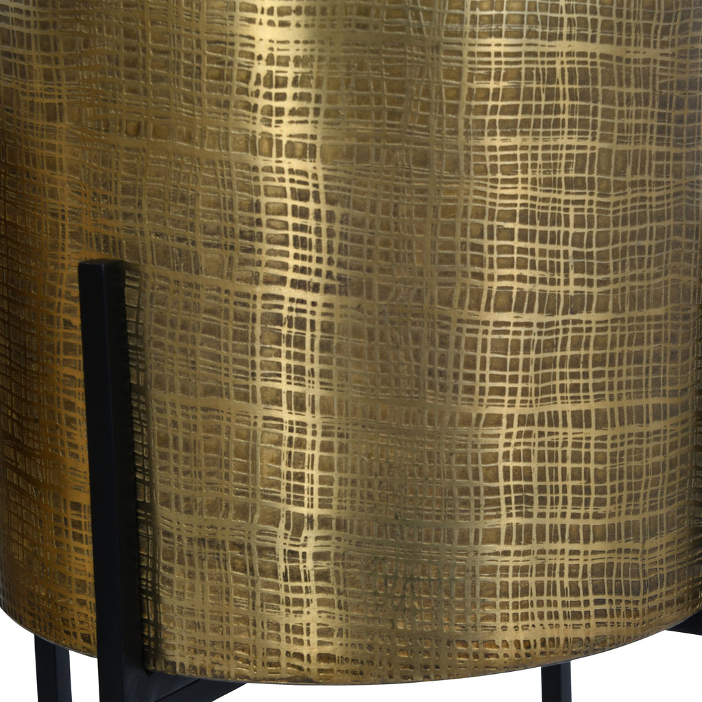 Detail of the etched rectangular pattern on a brass-coloured pot beside an upright black stand support.