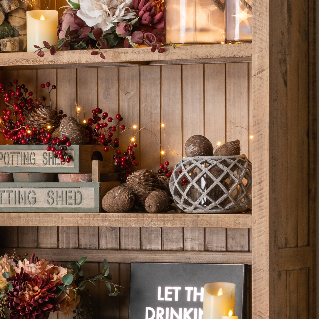 Crosslattice lantern displayed on a wooden shelf among pine cone decorations, red berry stems and warm string lights.