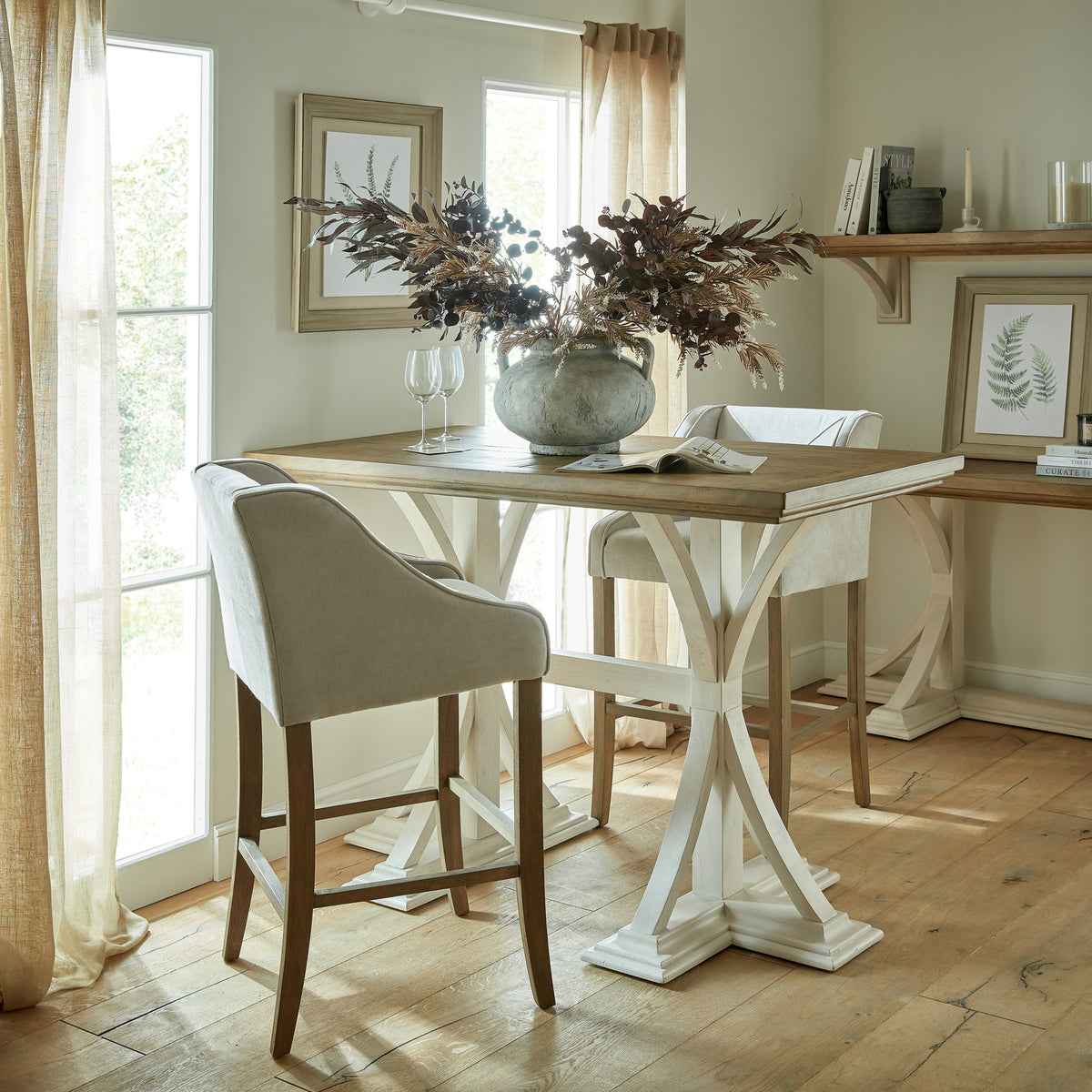 Two pale Crossstitch Counter Stools arranged around a tall table beside sunlit windows, with botanical prints and a large vase of foliage.