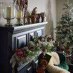 Red and burgundy berry picks among frosted foliage across a dark fireplace surround in a Christmas room with a decorated tree.