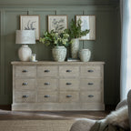 Pale-brown grid chest against a muted green panelled wall, styled with a lamp, patterned vases and foliage beneath three botanical prints.