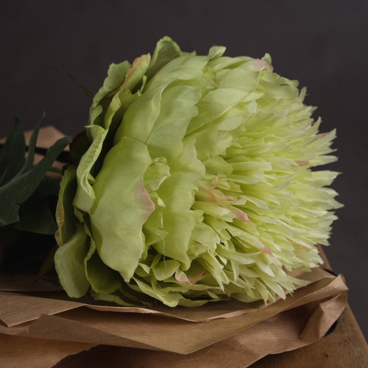 Side close-up of the flower head resting on brown paper, showing layered fabric petals with faint pink tips against a dark background.