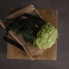 Overhead view of a single artificial flower stem with lobed green leaves on brown paper and black ribbon across a wooden stool.