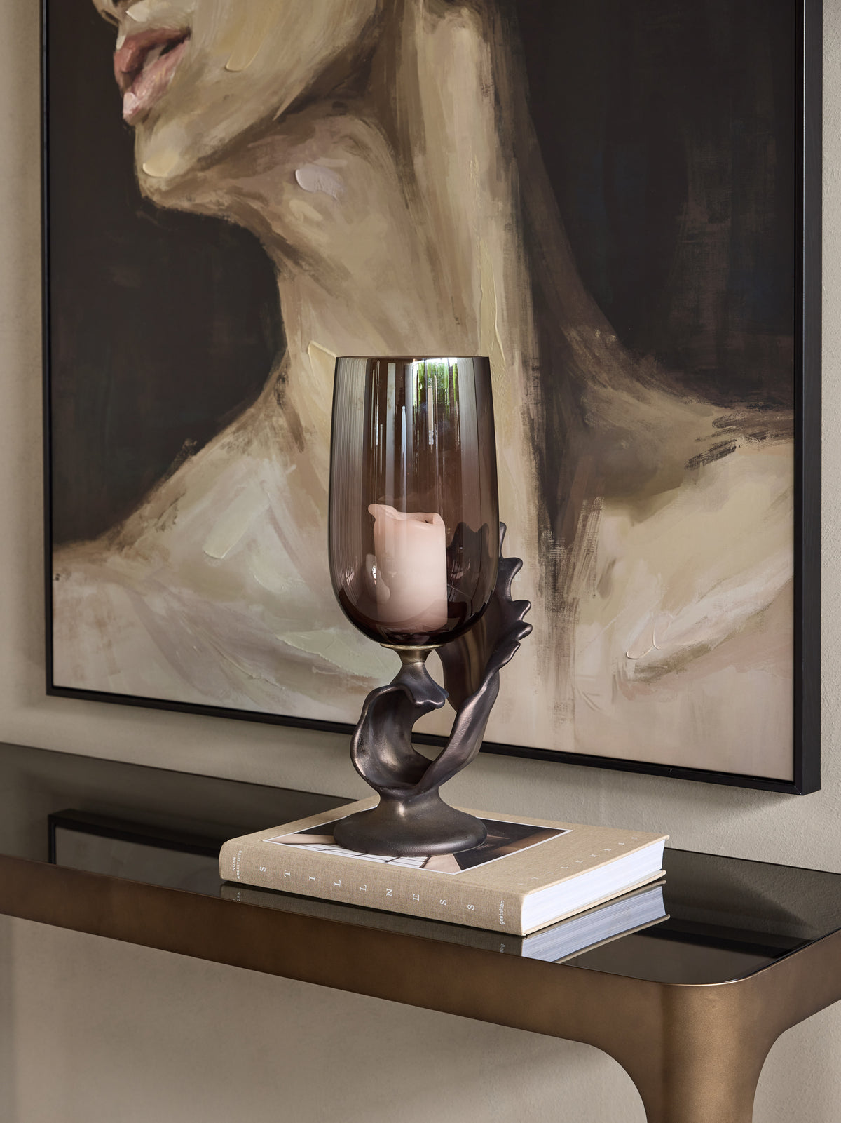 Hurricane candle holder containing a pale pillar candle, displayed on a book on a console beneath a large portrait.