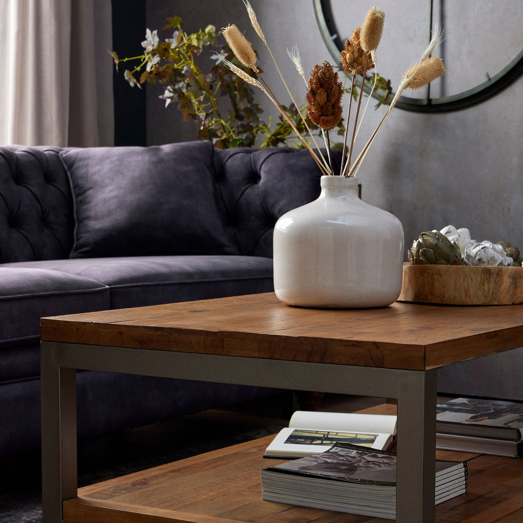 Cream botanical heads arranged with brown heads and fine pale stems in a white vase on a coffee table beside a dark sofa.