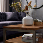 Cream botanical heads arranged with brown heads and fine pale stems in a white vase on a coffee table beside a dark sofa.