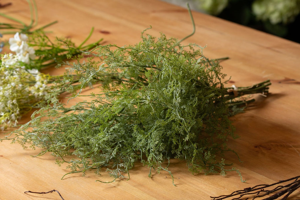 Curltip foliage laid across a wooden table, with tied stems pointing right and white flower stems beside the fine green leaves.