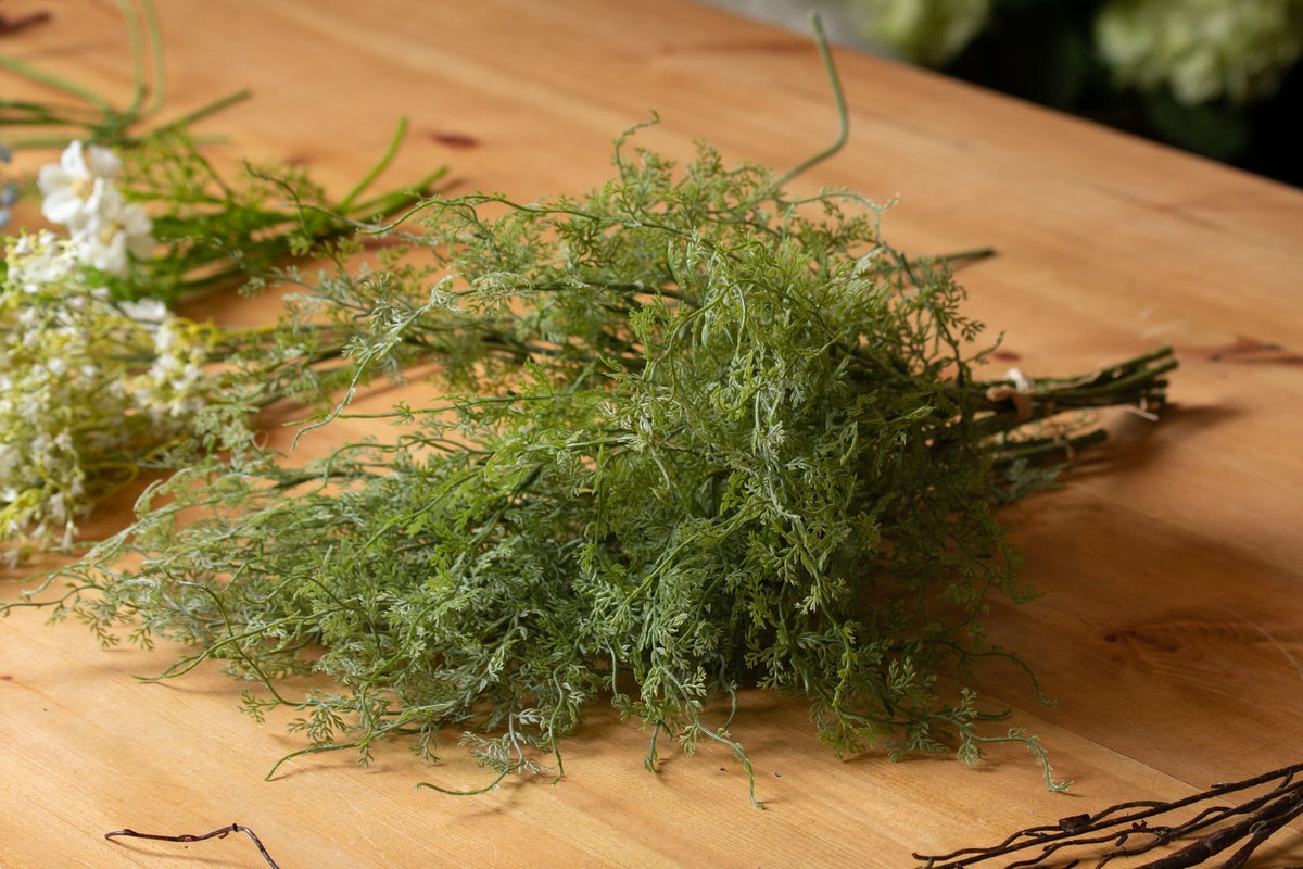 Curltip foliage laid across a wooden table, with tied stems pointing right and white flower stems beside the fine green leaves.