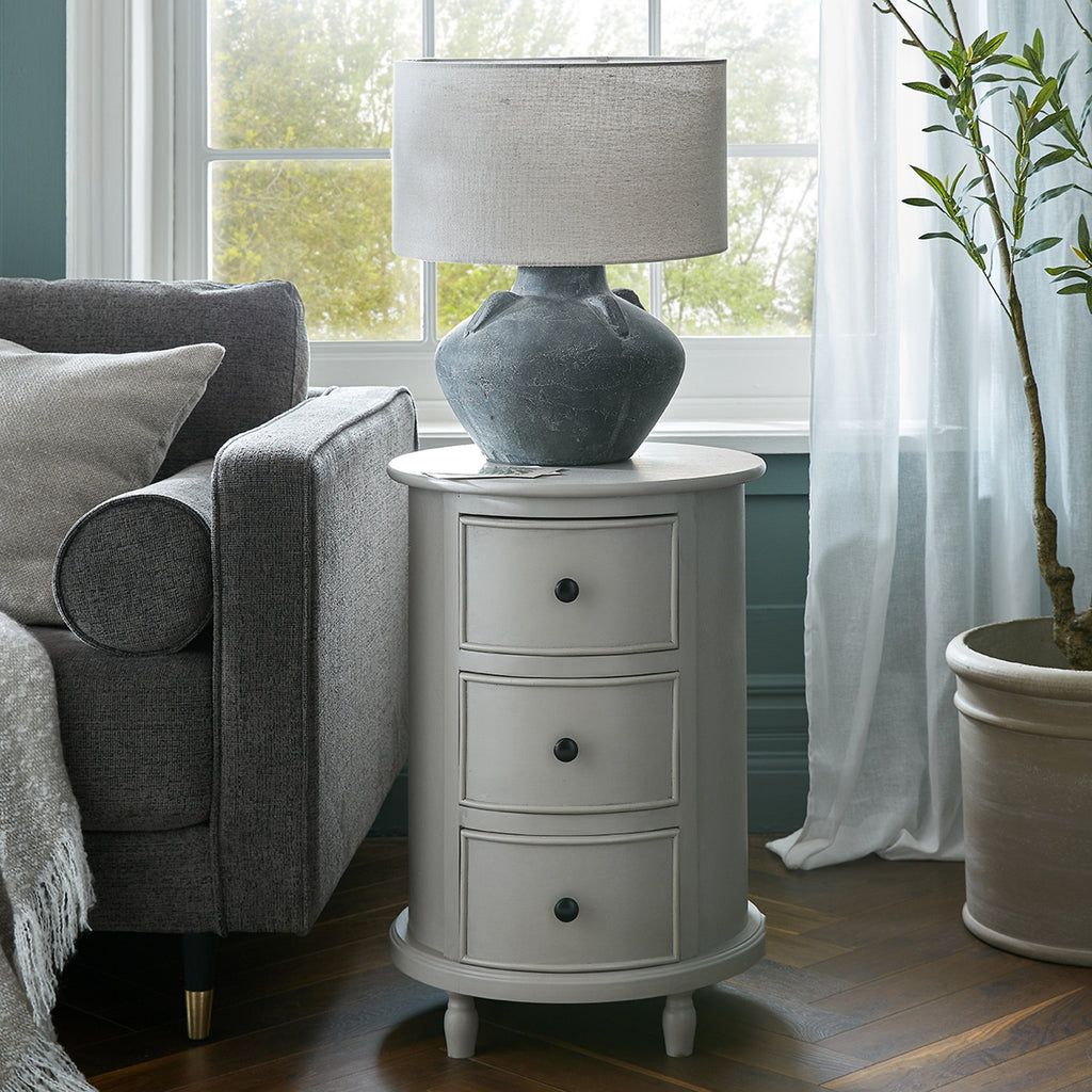 Round three-drawer side table beside a grey armchair, styled with a large grey lamp in front of a window, with pale curtains and a potted tree nearby.