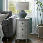 Round three-drawer side table beside a grey armchair, styled with a large grey lamp in front of a window, with pale curtains and a potted tree nearby.