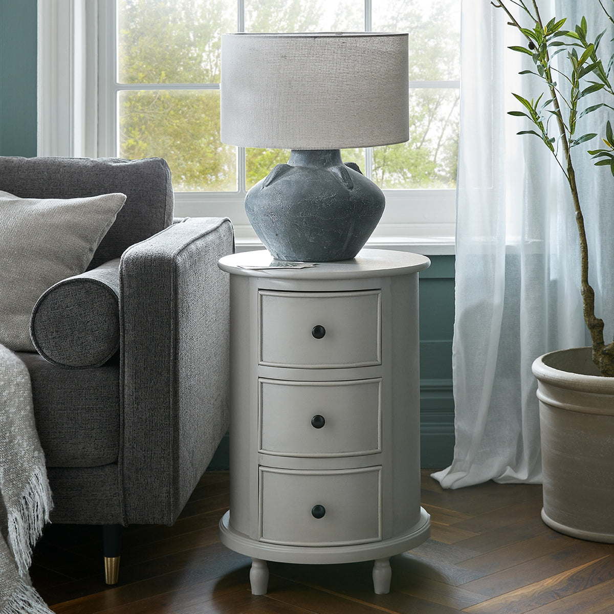 Round three-drawer side table beside a grey armchair, styled with a large grey lamp in front of a window, with pale curtains and a potted tree nearby.