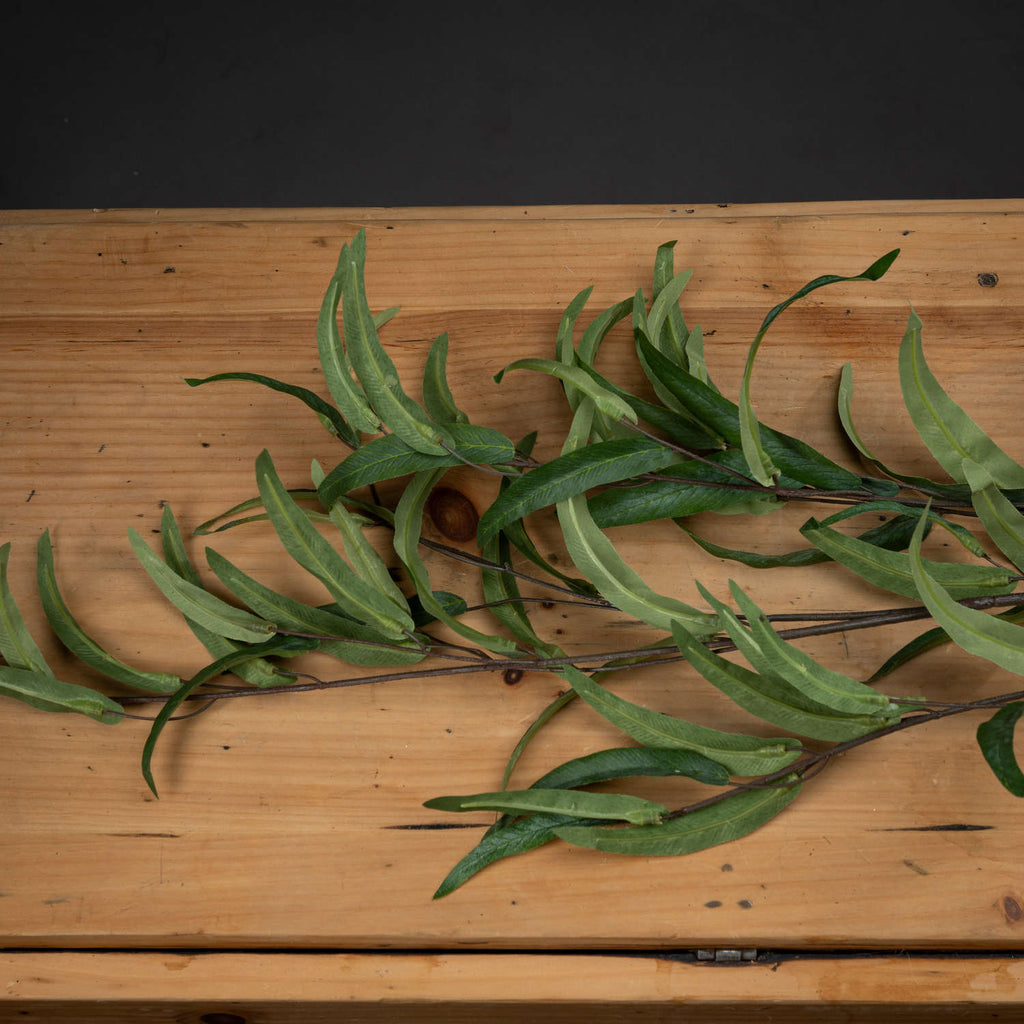 Artificial eucalyptus foliage laid across a wooden surface, showing narrow curling leaves with pale undersides and darker green upper faces.