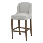 Front three-quarter view of a pale grey bar stool with a curved winged back, brown legs and rectangular lower rails.