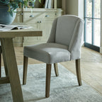 Pale grey curved-wing dining chair beside a timber-toned table on a patterned rug, with a cabinet and glazed doors behind.