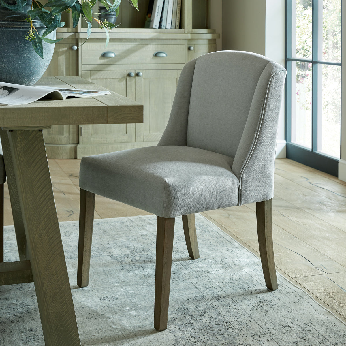 Pale grey curved-wing dining chair beside a timber-toned table on a patterned rug, with a cabinet and glazed doors behind.