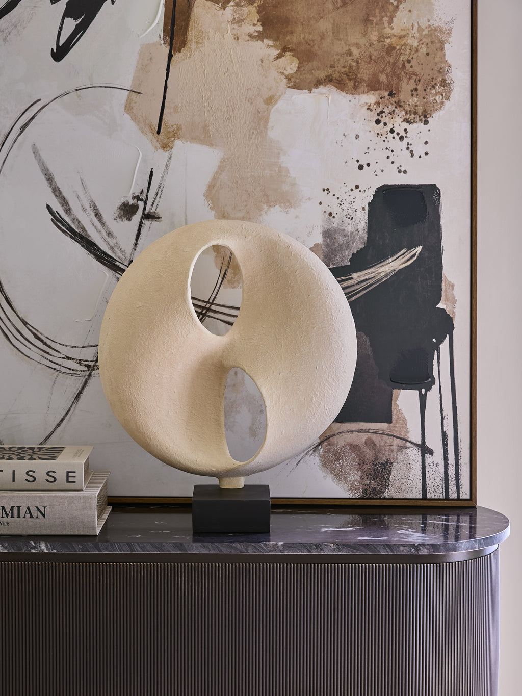 Cream cutout sculpture displayed beside stacked books on a dark console, with abstract artwork behind it.