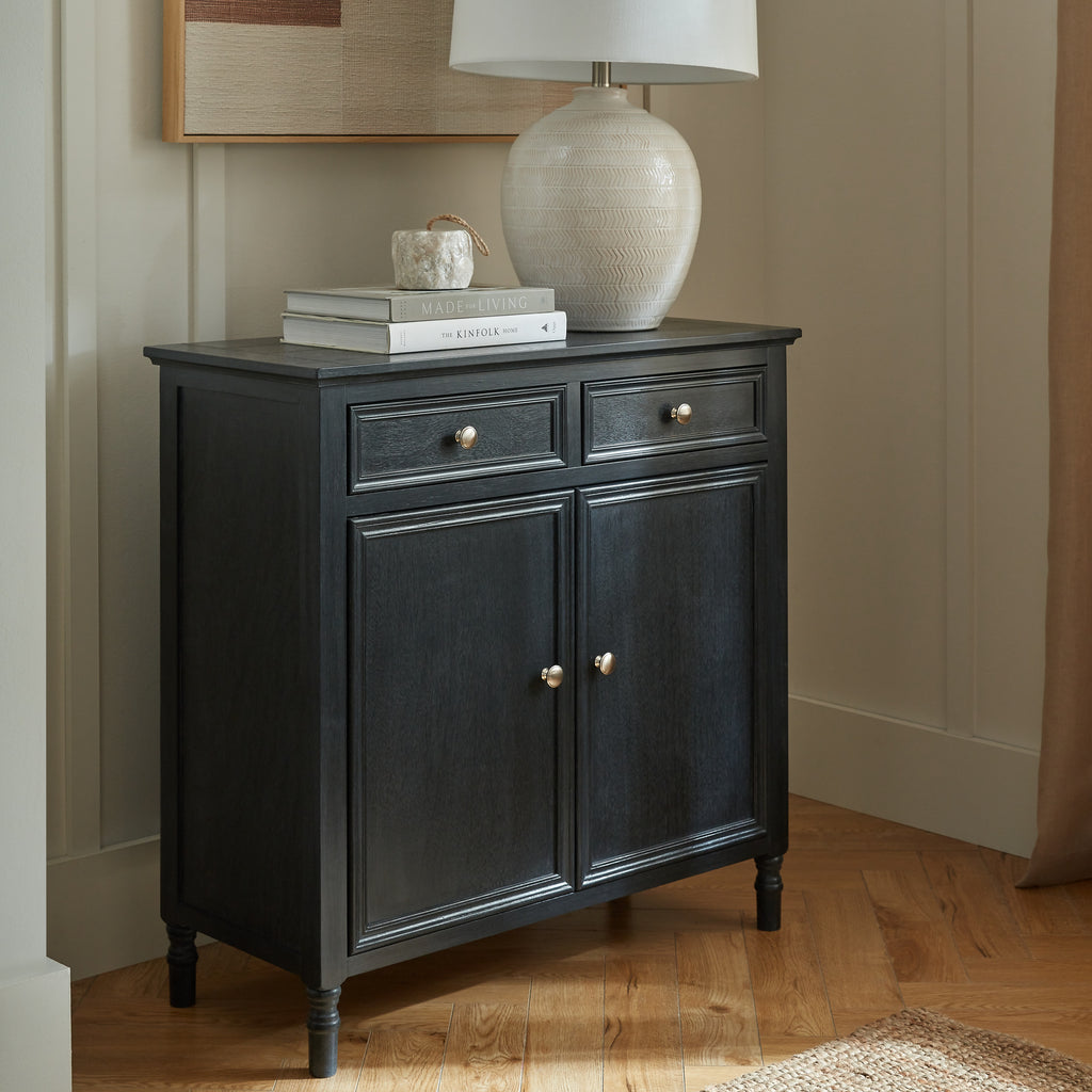 Dark panelled sideboard against a pale panelled wall, styled with books, a small decorative object and a cream table lamp beneath framed artwork.