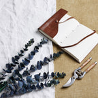 Dark foliage sprays arranged diagonally over white cloth beside a brown-covered open notebook and secateurs on a tan woven surface.