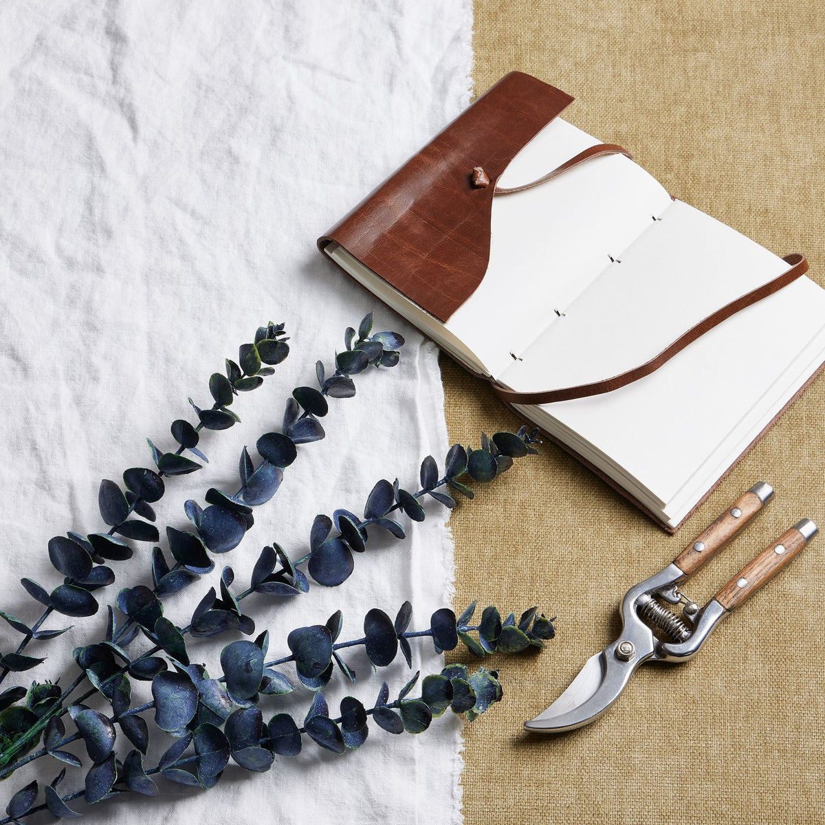 Dark foliage sprays arranged diagonally over white cloth beside a brown-covered open notebook and secateurs on a tan woven surface.