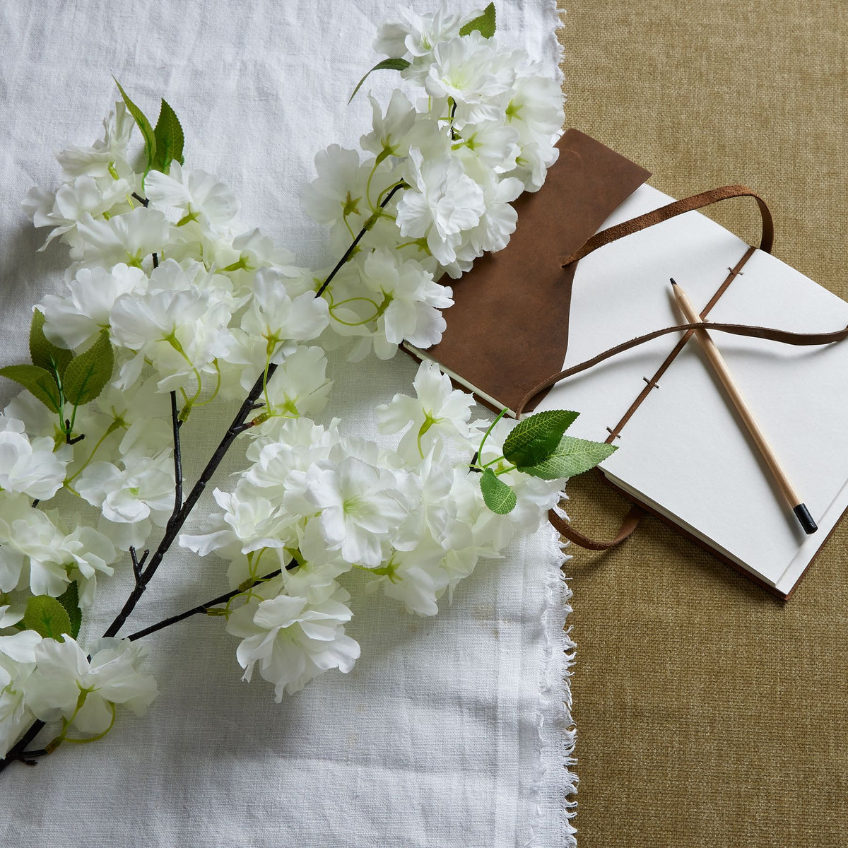 White blossom branches laid diagonally across white cloth beside an open brown notebook and pencil.