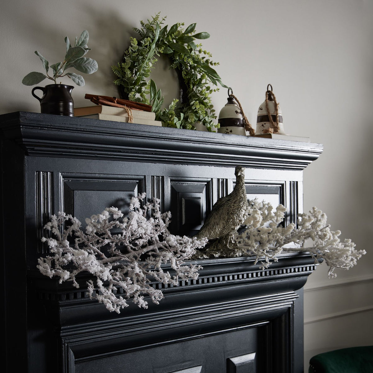 Snow-effect branches displayed across a dark fireplace surround around a bird ornament, below a green wreath, books and decorative vessels.
